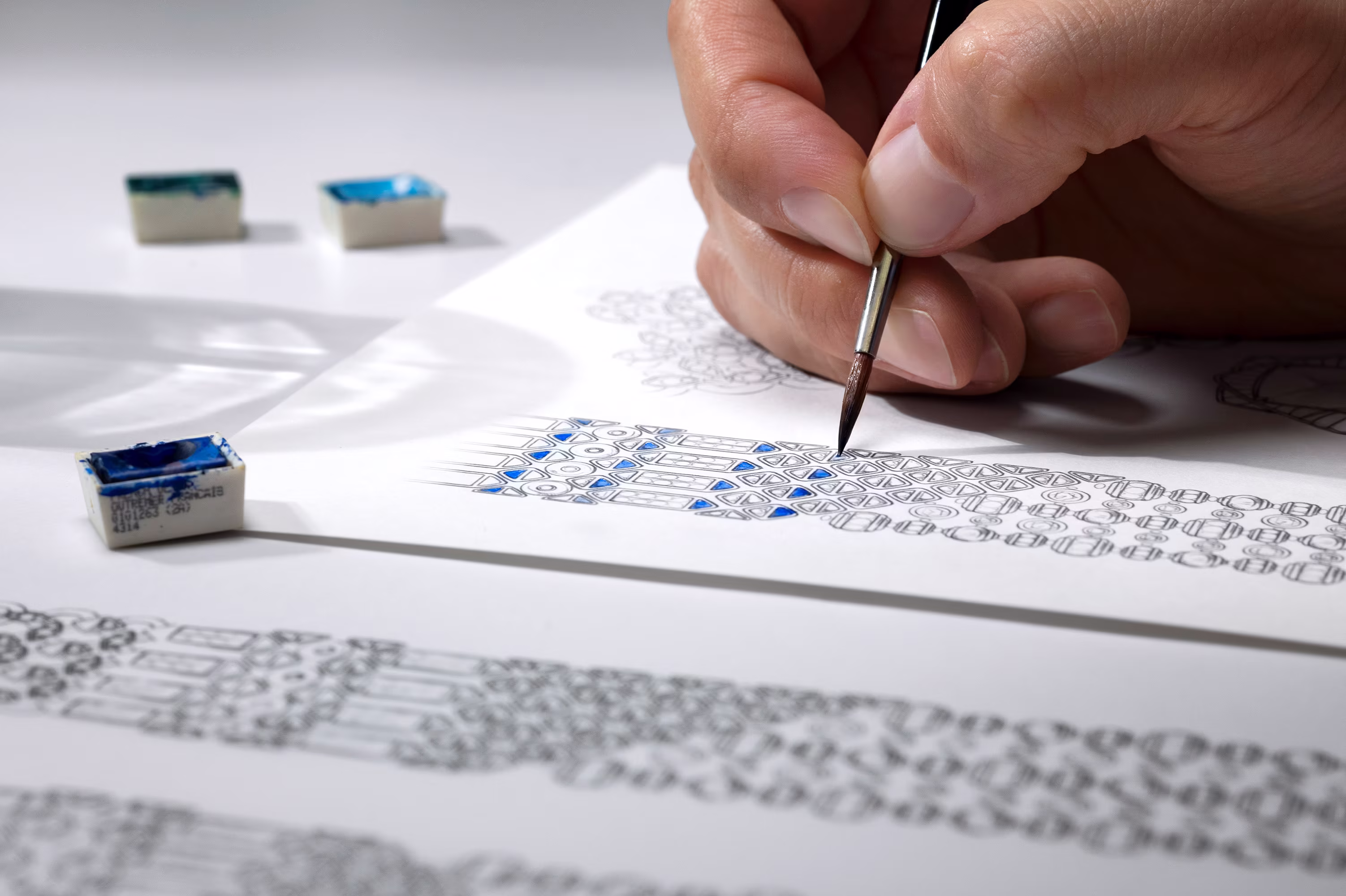 Hands paint a blue gemstone detail on a jewelry design sketch while holding a sapphire color sample nearby.